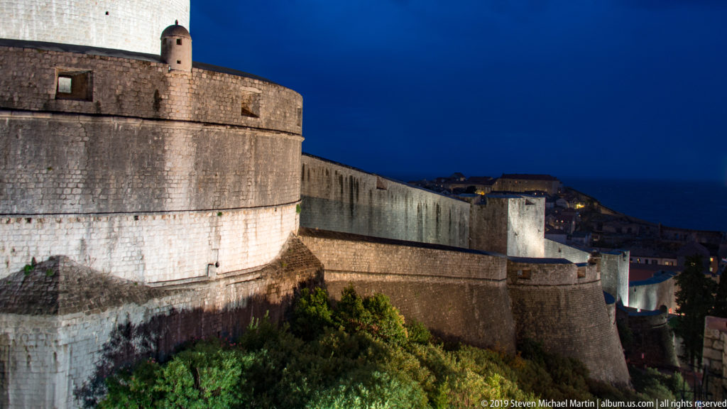 Rounded defensive tower and illuminated Dubrovnik city walls receding along the coast beneath a deep blue night sky.