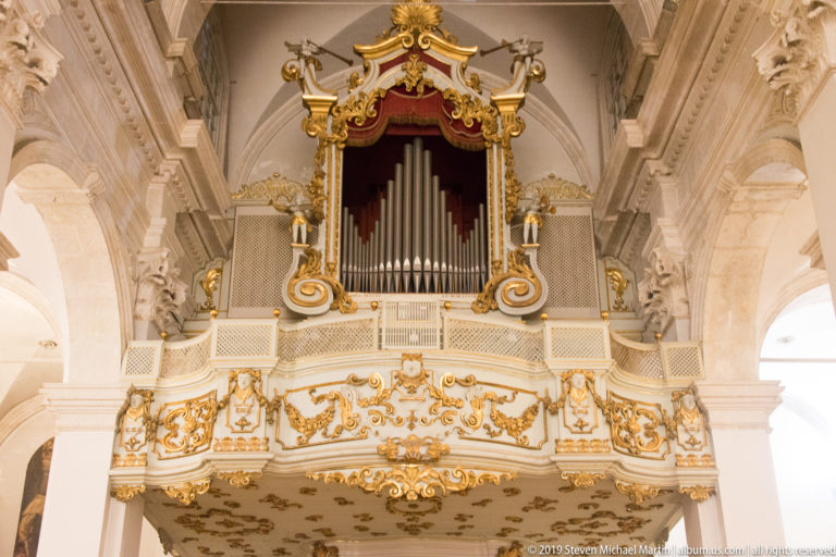 Ornate pipe organ above a white-and-gold gallery with red drapery, gilded scrollwork, and surrounding stone arches.