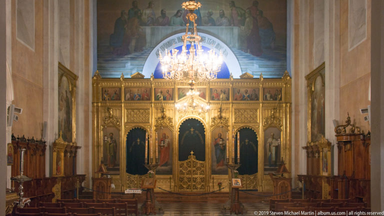 Orthodox church nave with chandelier, wooden seating, black-and-white floor, and gilded iconostasis at the sanctuary.