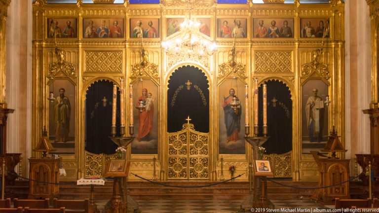 Frontal gilded Orthodox iconostasis with painted saints, pointed arches, carved tracery, royal doors, and candle stands.