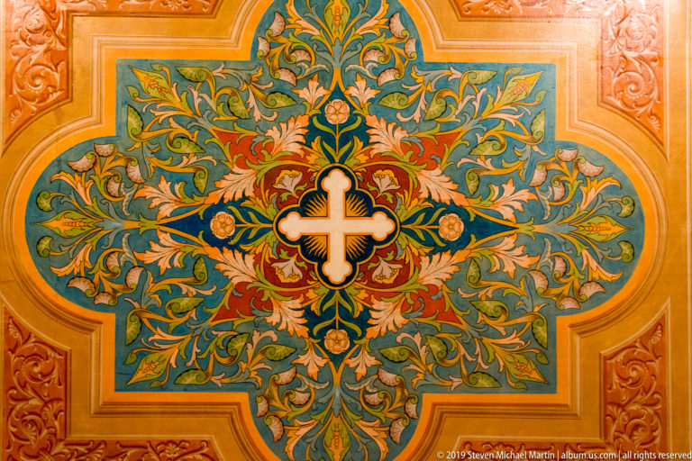 Polychrome ceiling ornament with central cross surrounded by symmetrical scrolling foliage, flowers, and molded framing.
