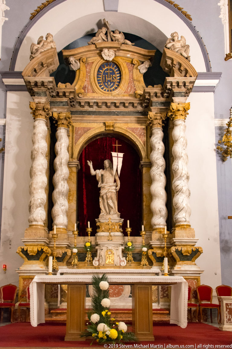 Baroque high altar with twisted columns, red niche, central statue, gilded ornament, flowers, and candles.