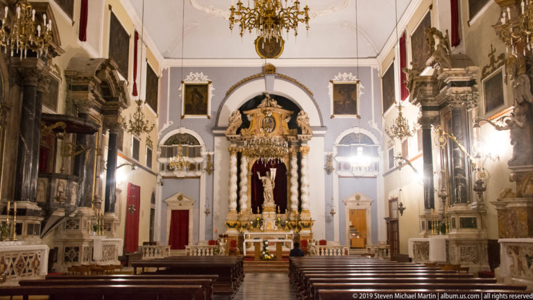 Symmetrical Baroque church nave with dark pews, side altars, chandeliers, paintings, and monumental high altar.