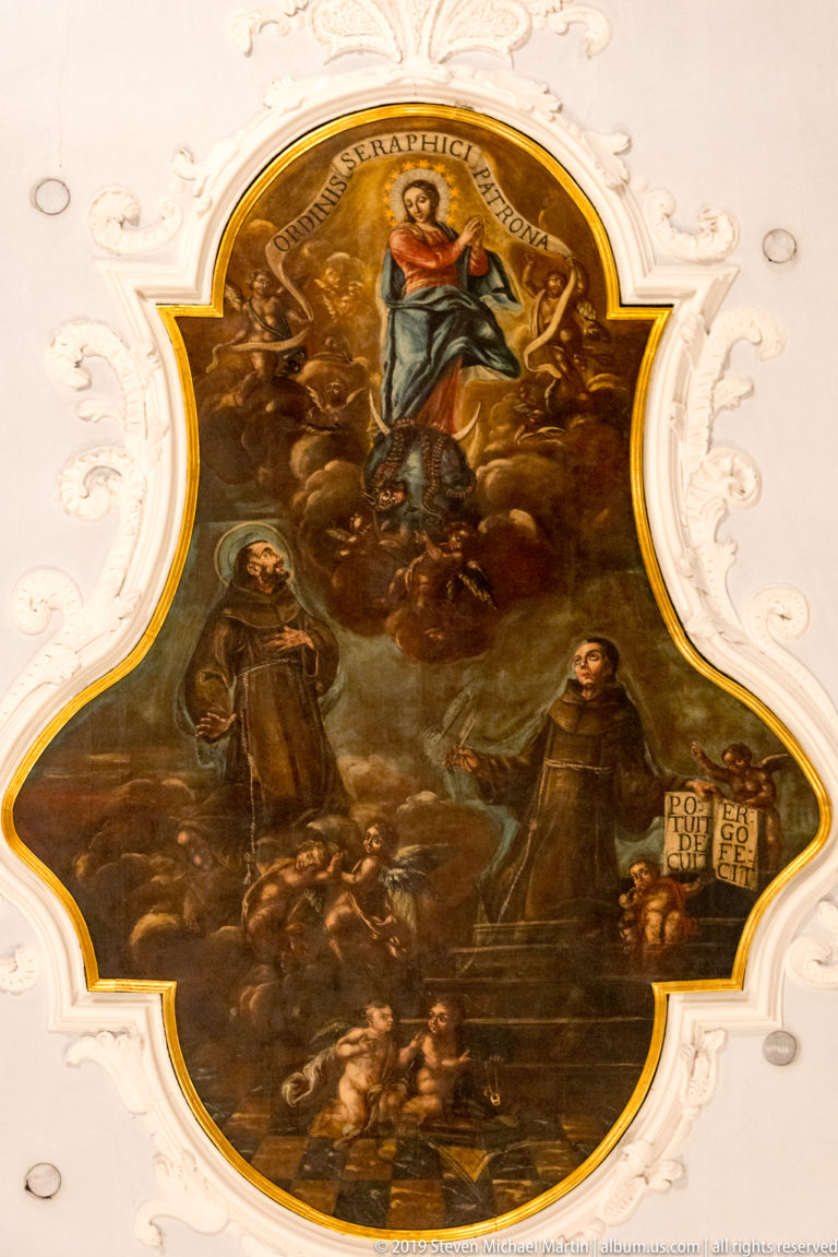 Baroque ceiling painting of a radiant holy figure above kneeling saints, framed by ornate white stucco decoration.
