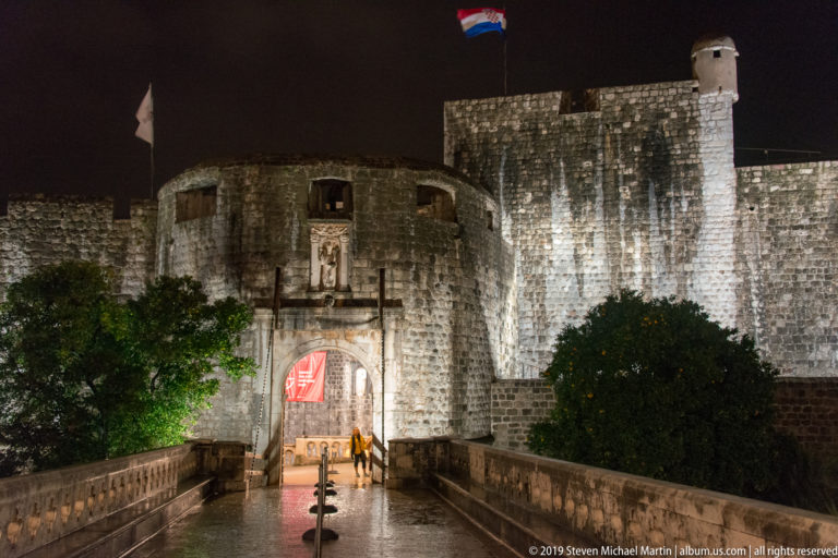 Outer Pile Gate with illuminated arch, sculpted niche, Croatian flag, bridge, and massive city walls at night.