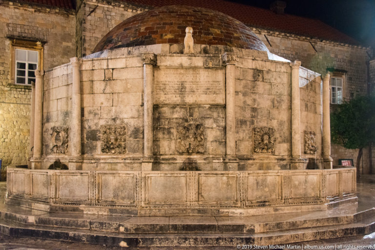Side view of the Great Onofrio Fountain showing polygonal stone basin, carved panels, water spouts, and low dome.
