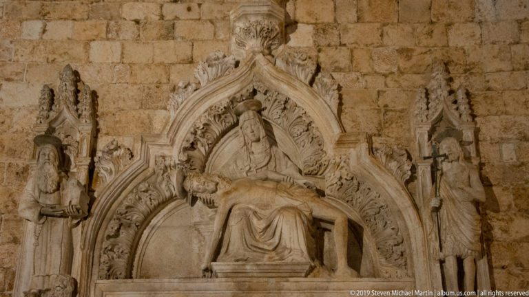 Stone Gothic relief of a seated Madonna and Child beneath a pointed canopy, flanked by two standing saints.