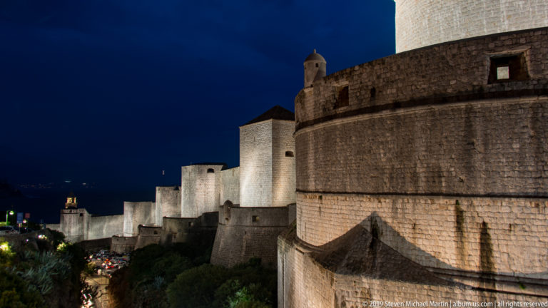 Illuminated eastern city walls of Dubrovnik with projecting towers and layered fortifications descending toward the harbor.