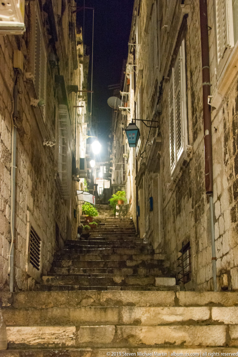 Steep stone steps climbing between close historic houses with shutters, signs, lamps, and plants in Dubrovnik at night.