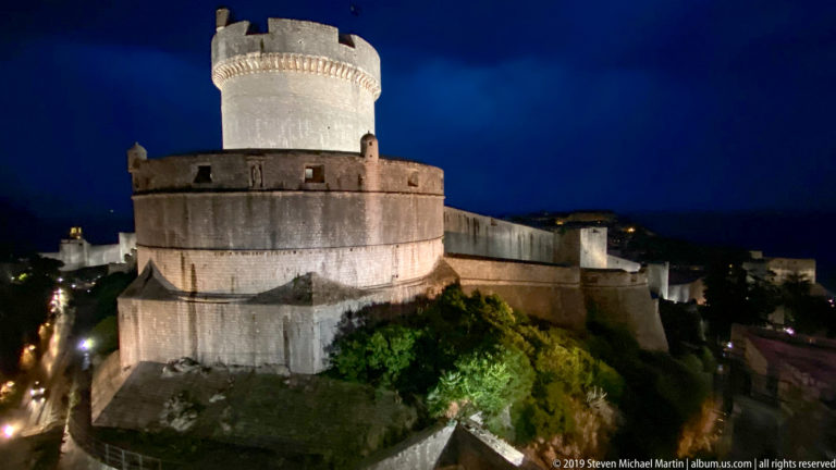 Oblique elevated night view of Minčeta Tower with circular masonry tiers and illuminated city walls extending behind.