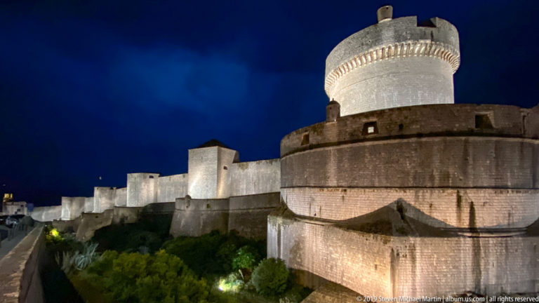 Illuminated Minčeta Tower with circular upper tower and long curtain walls beneath a deep blue night sky.