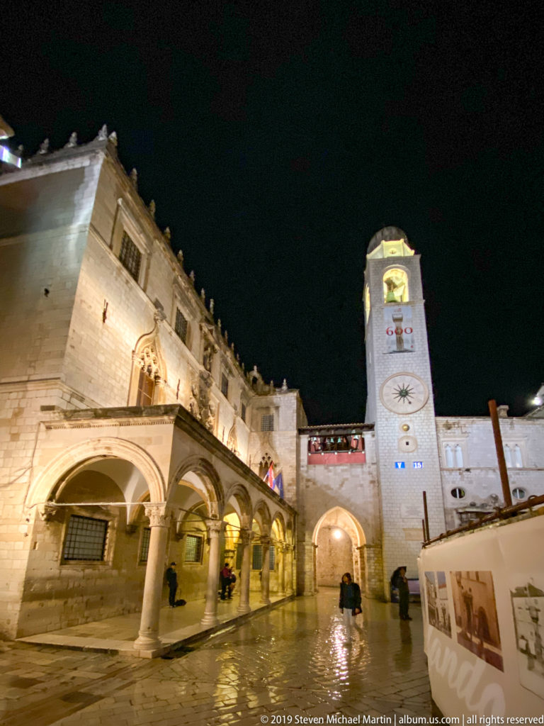 Wet Luža Square with Sponza Palace arcades at left and Dubrovnik City Bell Tower rising near the center.