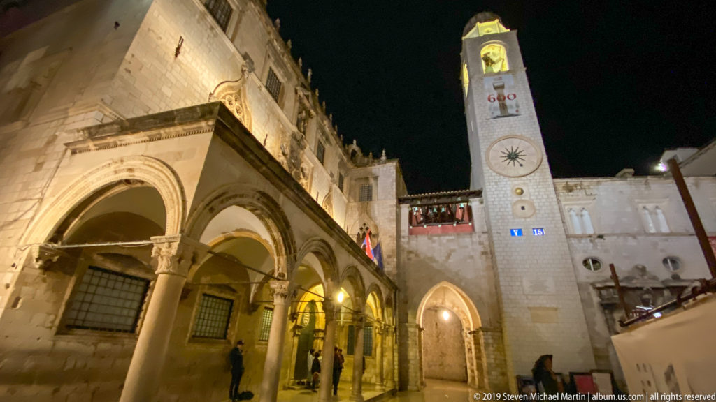 Low-angle view of Sponza Palace arcades beside Dubrovnik’s illuminated City Bell Tower at night.