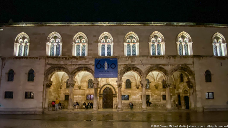 Illuminated Rector’s Palace with ground-floor arcade, Gothic upper windows, central doorway, and banner at night.