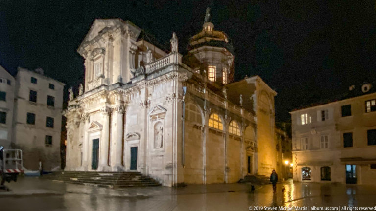Illuminated Church of St. Blaise with Baroque façade, broad steps, sculptures, and dome on wet Luža Square at night.