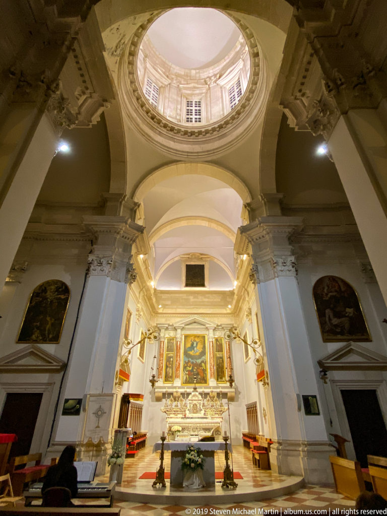 Baroque church interior with massive arches, circular dome, clerestory windows, paintings, chandeliers, and illuminated altar.