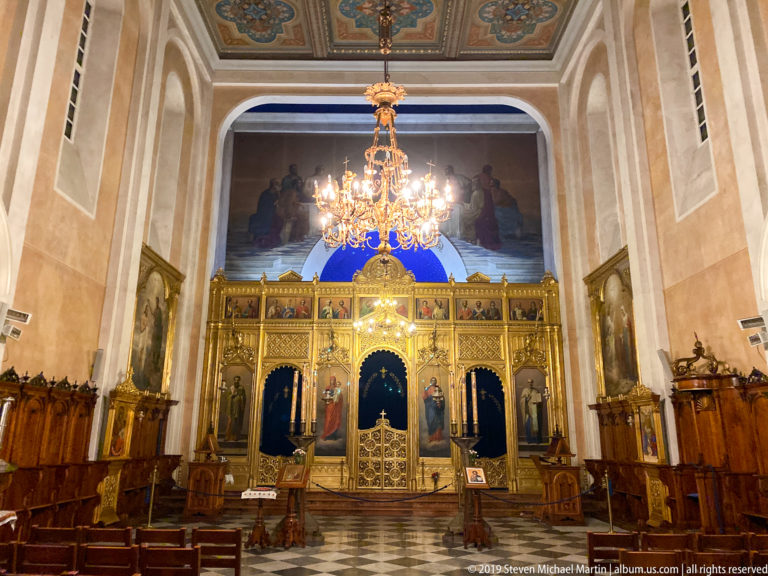 Frontal view of a gilded Orthodox iconostasis with painted icons, royal doors, chandelier, and wooden choir stalls.