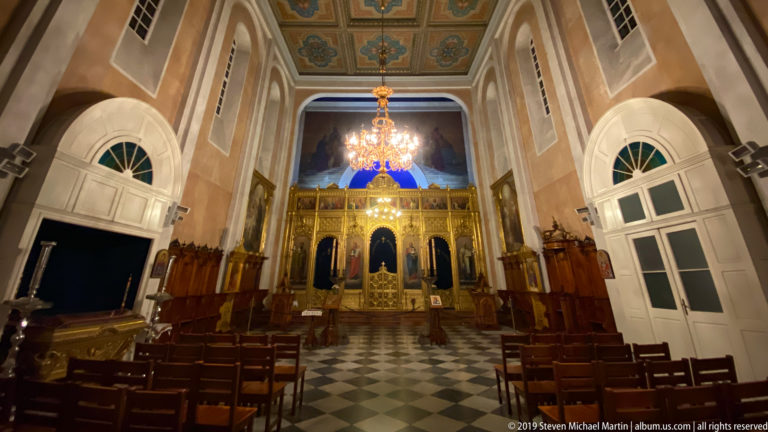 Church interior with gilded iconostasis, painted icons, chandelier, dark wooden stalls, and black-and-white tiled floor.