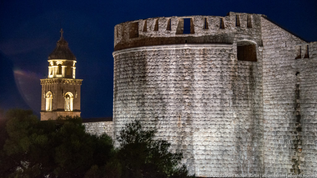Illuminated rounded defensive tower on Dubrovnik’s city walls with a lit church bell tower rising behind at night.