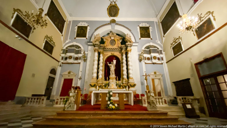 Baroque church altar with marble columns, red niche, central statue, flowers, candles, and gilded architectural ornament.
