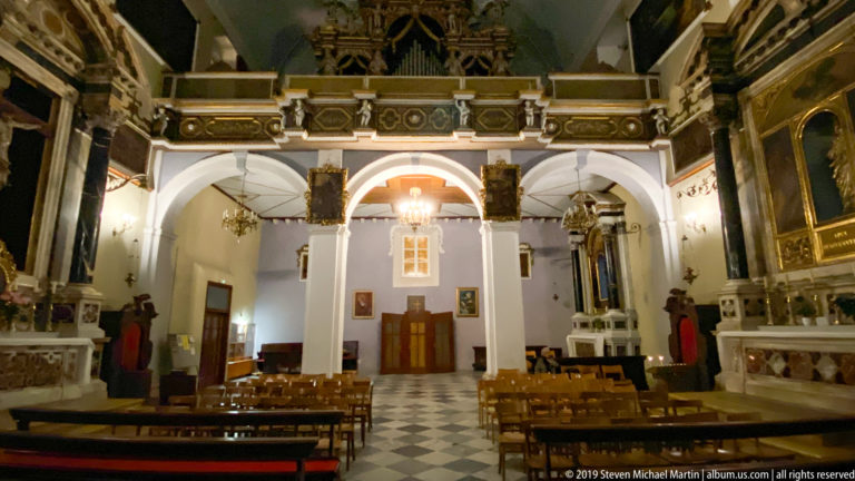 Historic church interior with black-and-white floor, arcades, side altars, chandeliers, paintings, and rows of chairs.