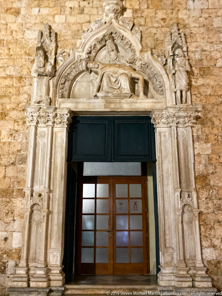 Pointed Gothic church portal with sculpted figures, tracery, and tympanum set into a rough stone wall at night.