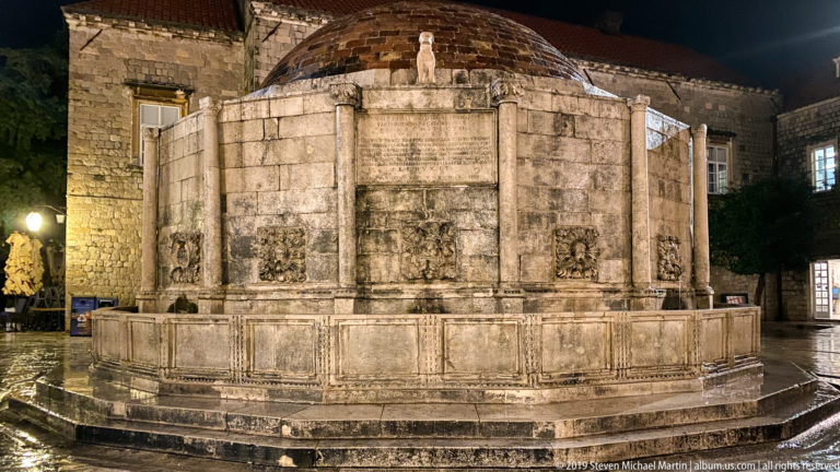 Great Onofrio Fountain at night, showing its polygonal stone basin, relief carvings, spouts, and low domed roof.