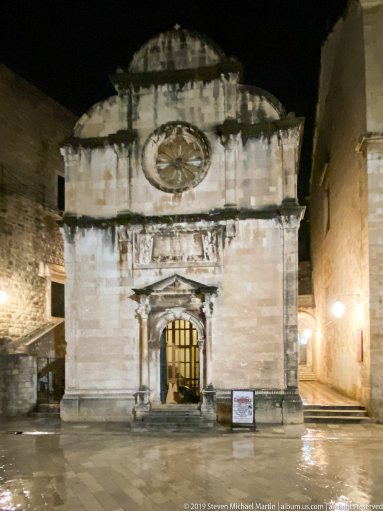 Illuminated façade of St. Saviour Church with rose window, sculpted portal, and curved gable on wet paving at night.