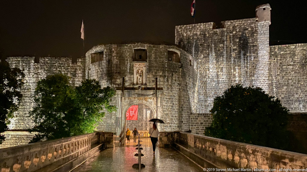 Rainy nighttime approach to outer Pile Gate with pedestrians, sculpted niche, massive walls, and flag above.