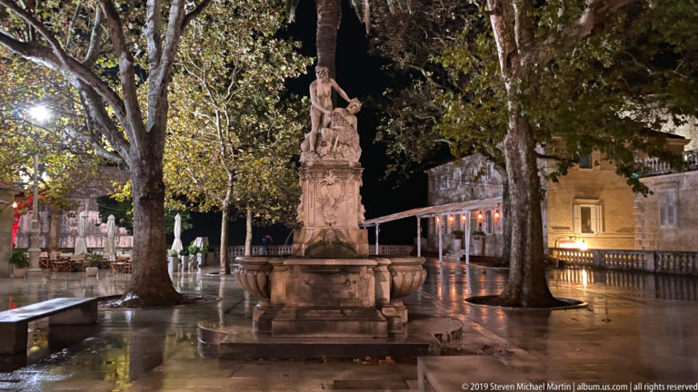 Decorative stone fountain with standing figure beneath trees in a wet square near Dubrovnik’s Pile entrance at night.