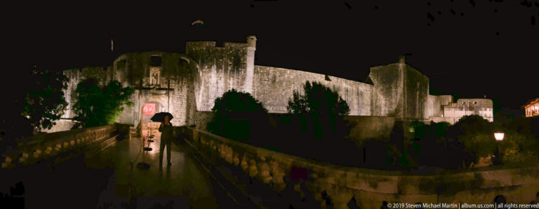 Wet bridge leading toward Pile Gate beneath illuminated western city walls and a Croatian flag at night.