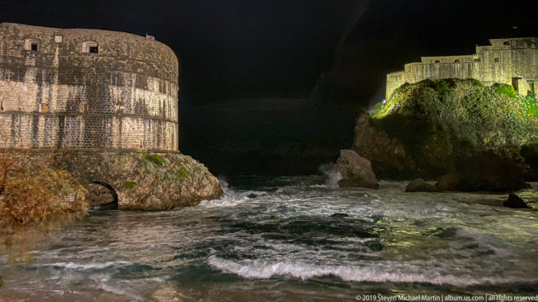 Night surf between rounded Bokar Fortress at left and illuminated Lovrijenac Fortress on the rocky outcrop at right.