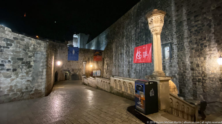 Wet inner Pile Gate walkway with carved column, stone balustrade, banners, and high masonry walls at night.
