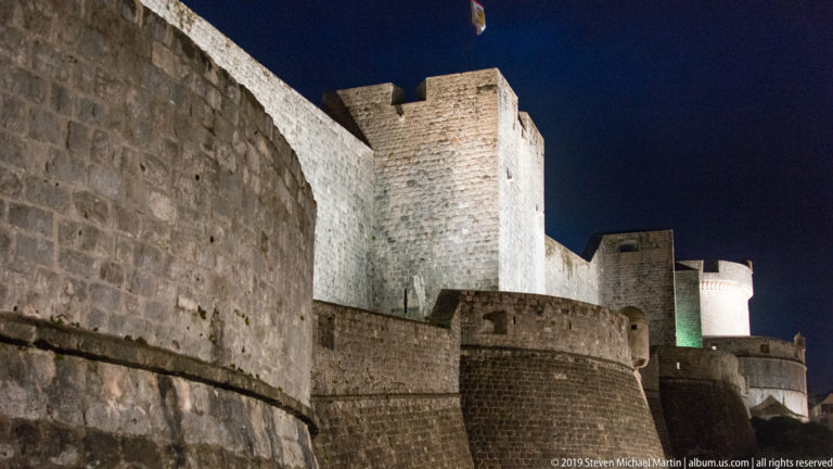 Illuminated Dubrovnik city walls and towers rising in successive stone layers beneath a dark blue night sky.