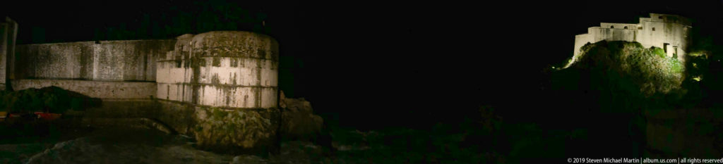 Rounded Bokar Fortress in front and illuminated Lovrijenac Fortress on the rocky promontory beyond at night.