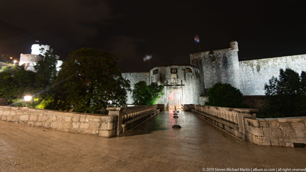 Night view of the bridge to Pile Gate beneath illuminated curtain walls and higher fortifications at Dubrovnik’s western entrance.