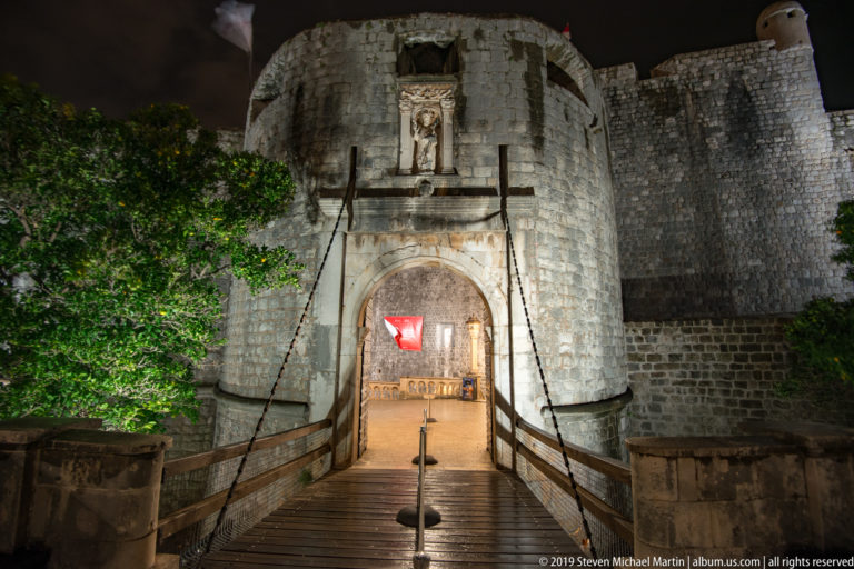Outer Pile Gate centered within massive illuminated city walls, with a wet stone bridge leading to the entrance.