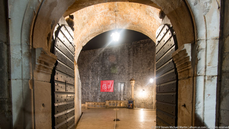 Open studded wooden doors beneath a stone arch framing the illuminated inner passage of Dubrovnik’s Pile Gate.