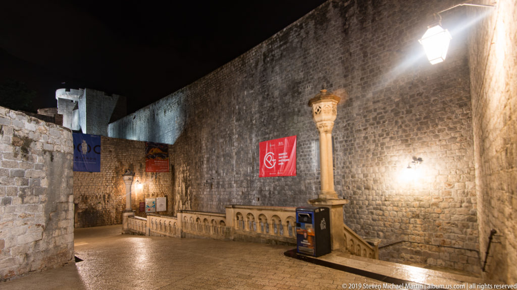 Wet stone passage with balustrade, banners, carved column, and high defensive walls at Dubrovnik’s western gate approach.