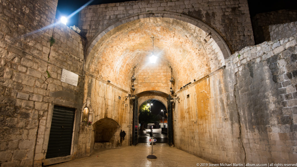 Barrel-vaulted stone gate passage with lamps, side doors, and a dark opening through Dubrovnik’s fortifications.