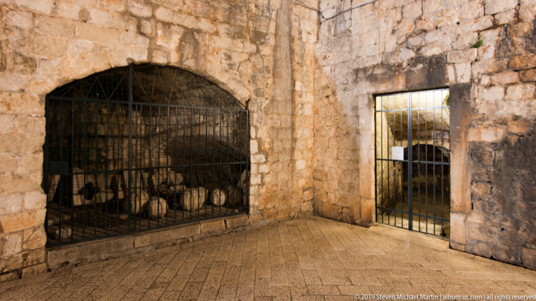 Heavy iron grilles closing stone recesses with stored vessels or equipment visible behind one gate.