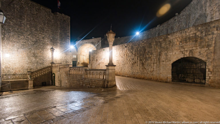 Stone-paved fortified courtyard with ramp, balustraded stair, high walls, and arched gate opening at night.