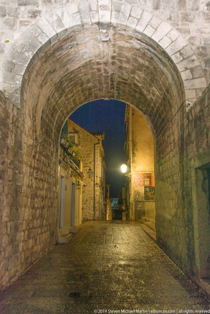 Deep stone archway through Dubrovnik’s fortifications, with wet pavement and a quiet street visible beyond at night.