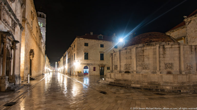 Great Onofrio Fountain beside wet Stradun and illuminated historic buildings near Dubrovnik’s Pile entrance at night.