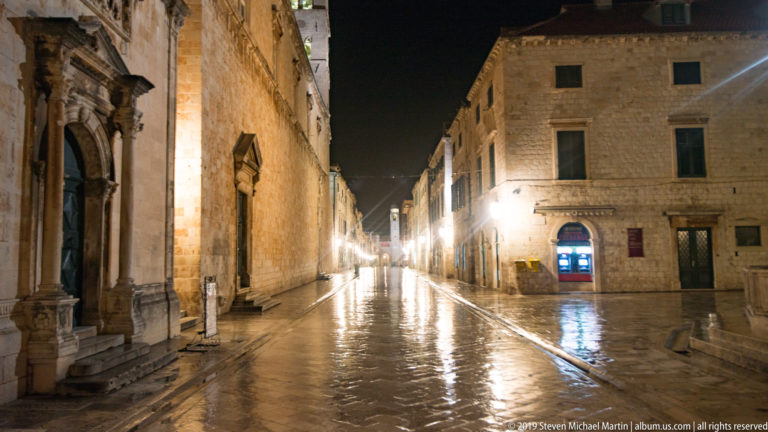 Wet limestone street junction lined by historic stone buildings and a carved church portal in Dubrovnik at night.