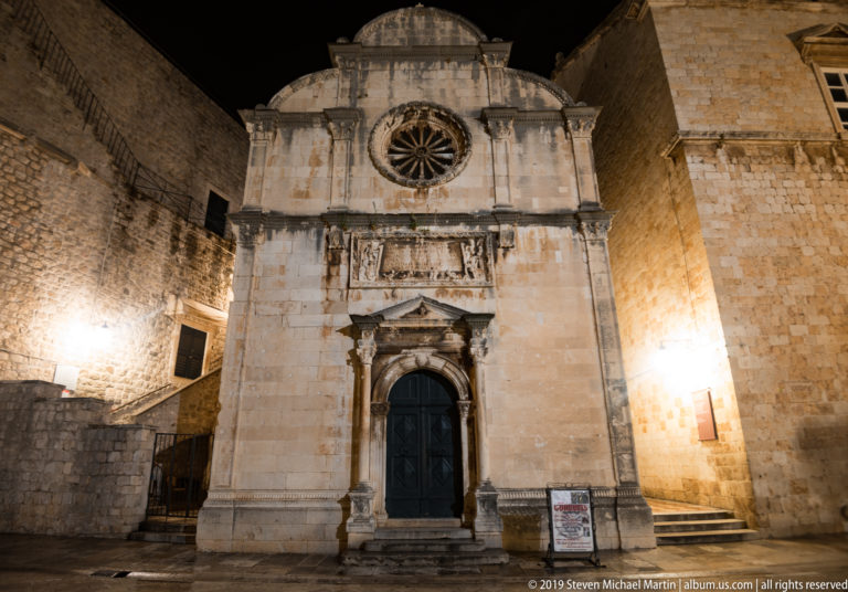 Frontal illuminated façade of St. Saviour Church with rose window, carved portal, and city walls behind.