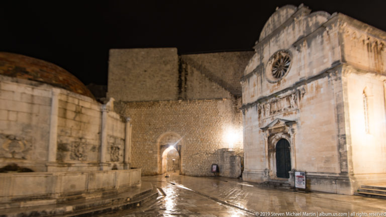St. Saviour Church with rose window and sculpted portal beside massive stone walls and an arched passage at night.