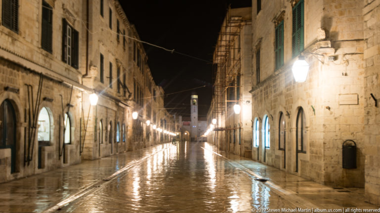 Centered nighttime view along wet Stradun between tall stone façades and evenly spaced street lamps.