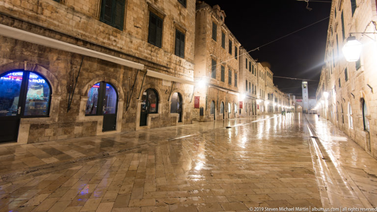 Long nighttime view down wet Stradun with arcaded stone façades, shopfronts, and repeating street lamps.