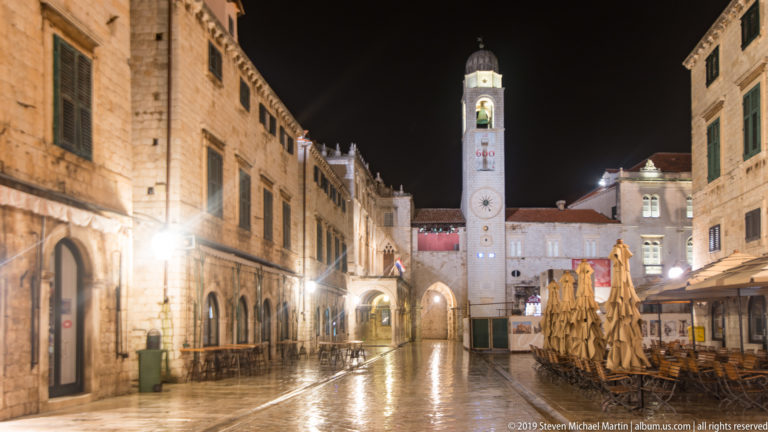 Wet pedestrian street lined by stone façades and closed café umbrellas leading toward the City Bell Tower at night.